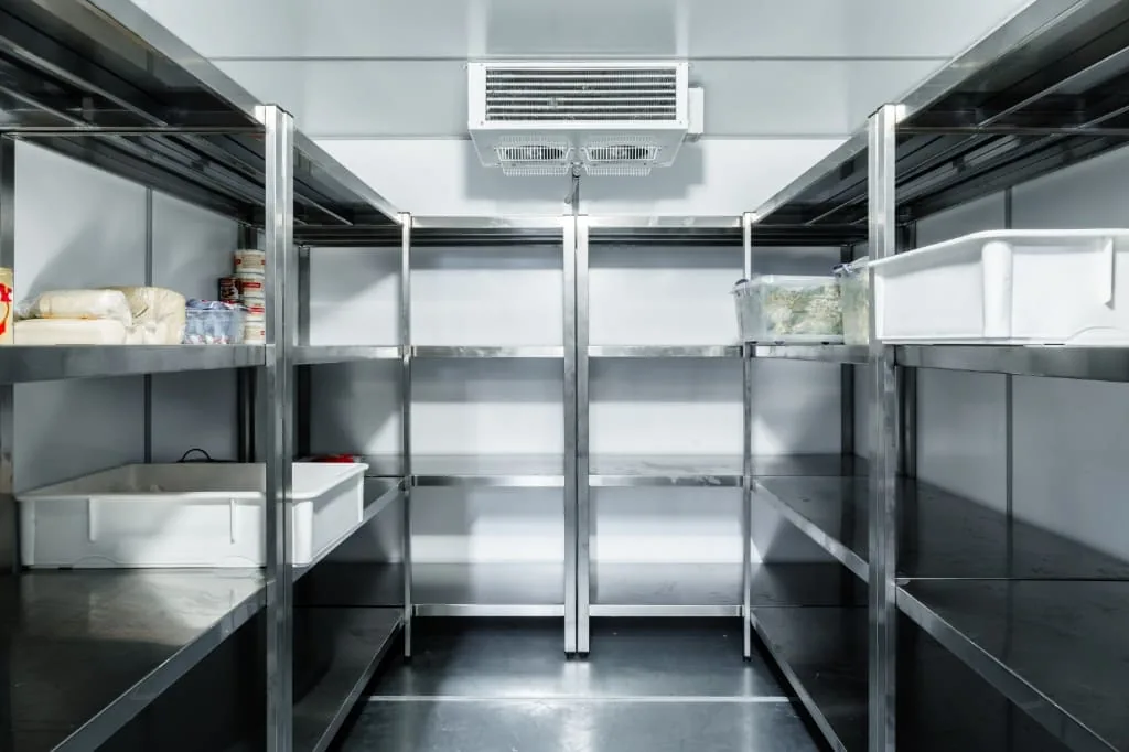 Interior de una cámara frigorífica con estanterías de acero inoxidable, cubetas blancas y el evaporador en el techo