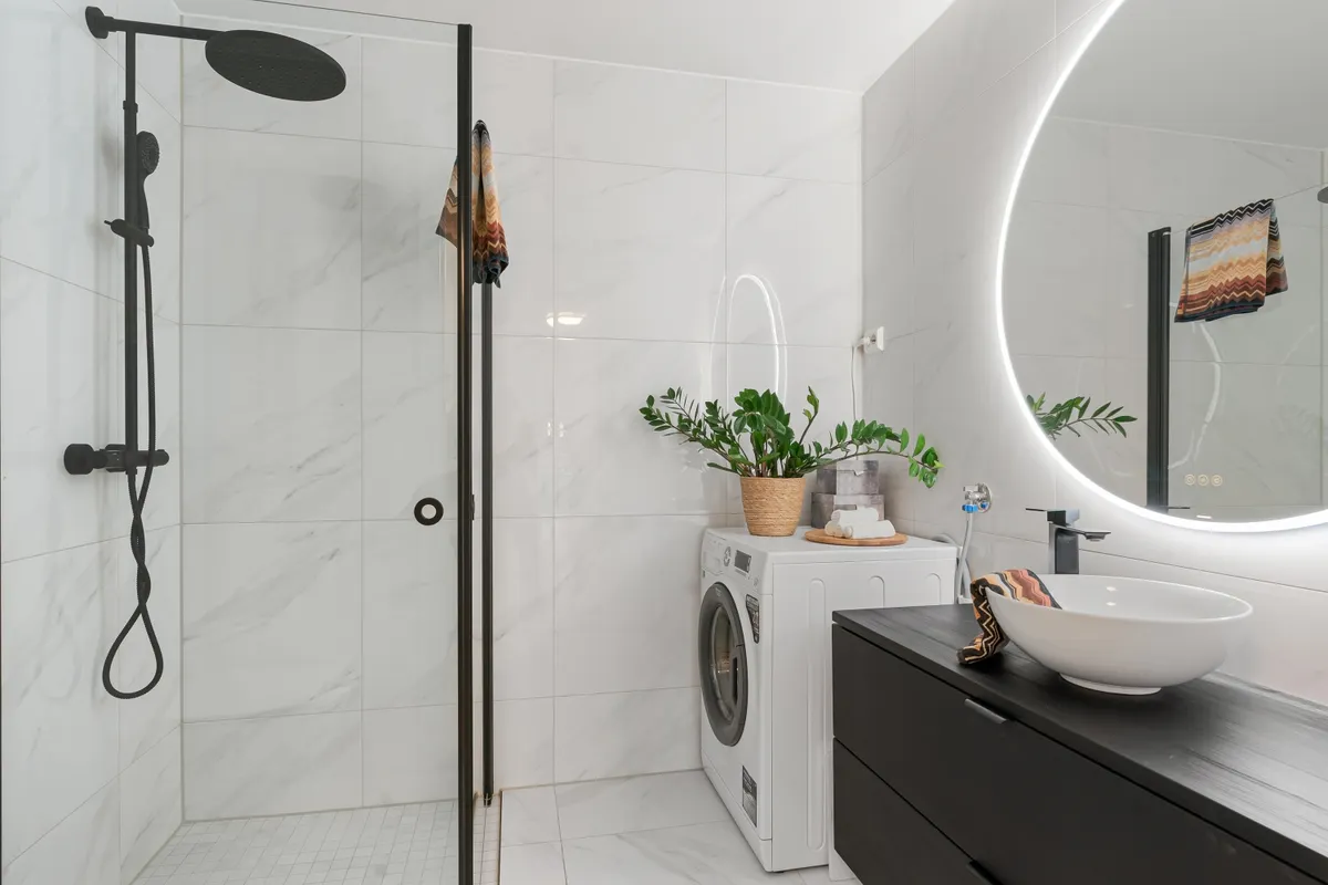 Baño moderno con una lavadora de carga frontal entre la ducha y el lavabo, con la toma de agua y el enchufe en la pared