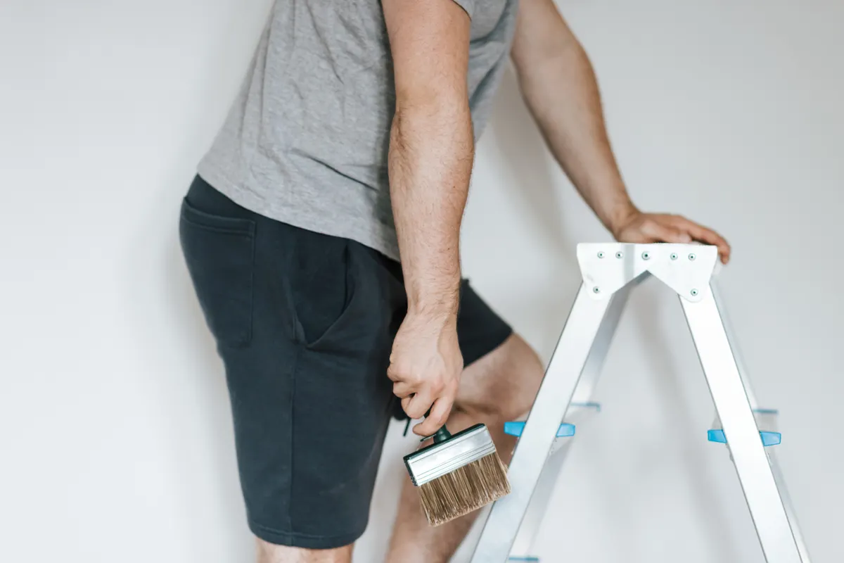 Pintor con ropa informal subido a una escalera de tijera preparándose para pintar una habitación luminosa